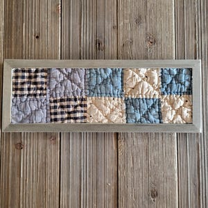 May include: A rectangular wall art piece featuring a quilted fabric panel with a grid of ten squares in blue, cream, and black-and-white gingham patterns, all encased within a simple, slim, metallic silver-toned frame for display.