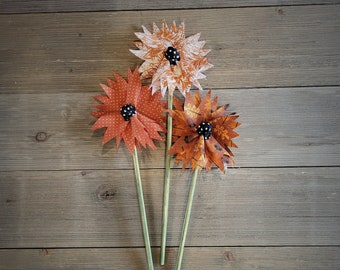 May include: Three paper flowers with orange, brown, and white patterned fabric. The flowers have black button centers and are attached to green stems.