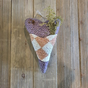 May include: A purple and pink heart-shaped fabric cone with a dried flower arrangement inside. The cone is hanging on a wooden wall.