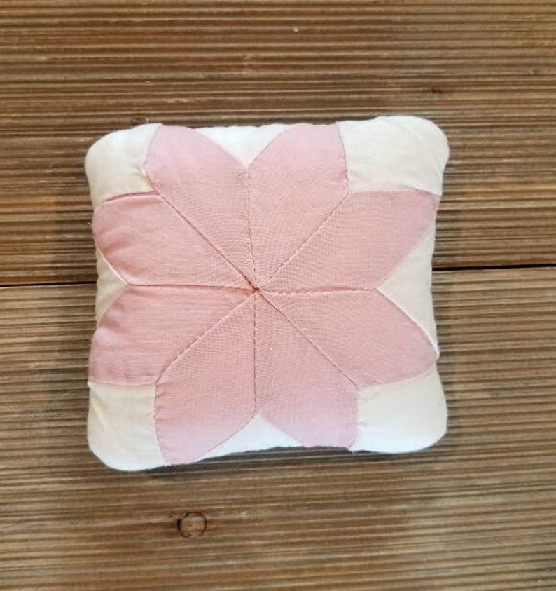 May include: A pink and white patchwork pillow with a starburst design. The pillow is made of soft fabric and has a subtle sheen.