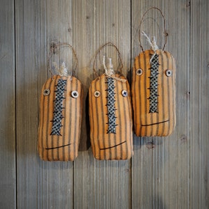 May include: Three handmade orange and black striped fabric pumpkins with stitched faces. The pumpkins have black eyes and mouths and are hanging from a wooden wall.