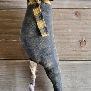 May include: A handmade, dark gray fabric bird with a yellow and black plaid bow. The bird is mounted on a small wooden stick. The bird has a distressed, vintage appearance, and is likely a decorative item.