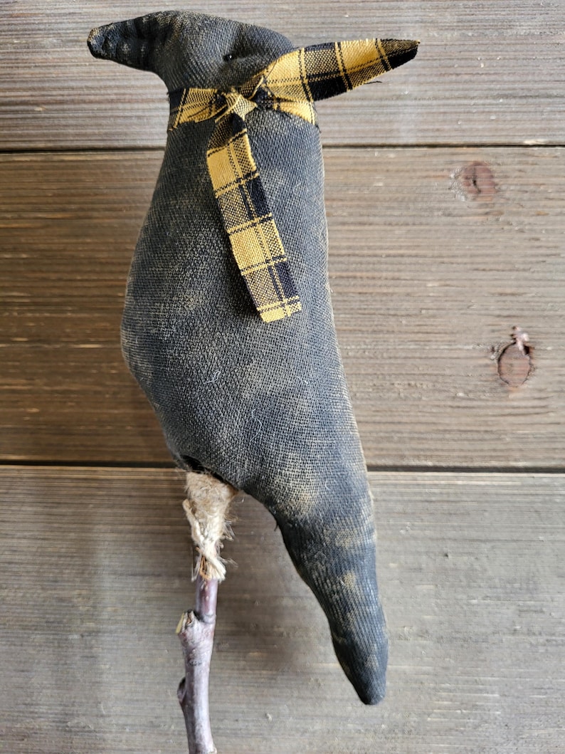 May include: A handmade, dark gray fabric bird with a yellow and black plaid bow. The bird is mounted on a small wooden stick. The bird has a distressed, vintage appearance, and is likely a decorative item.