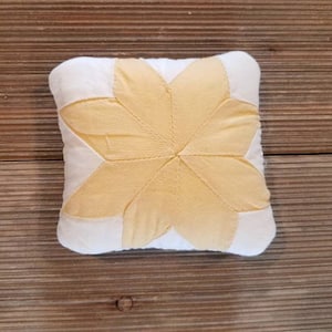 May include: A small, white pillow with a yellow star-shaped design in the center. The pillow is made of fabric and has a soft, quilted texture.
