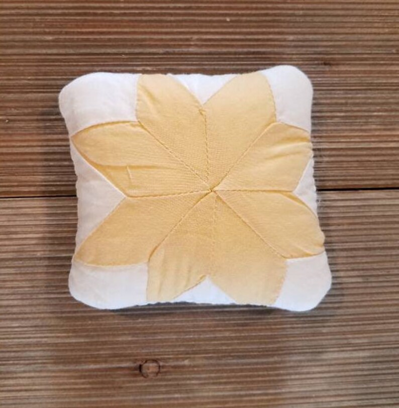 May include: A small, white pillow with a yellow star-shaped design in the center. The pillow is made of fabric and has a soft, quilted texture.