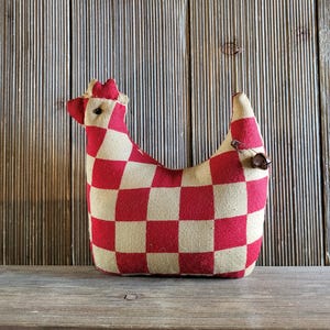 May include: A stuffed fabric chicken ornament featuring a red and beige checkered pattern. The chicken has a small red comb on its head, a dark bead eye, and a metal safety pin with a small dangling bell attached to its tail section.