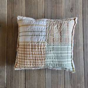May include: Square patchwork pillow with a variety of patterns and colors. The pillow features sections of white, tan, and green stripes and plaid. The pillow has a quilted design and a red trim.
