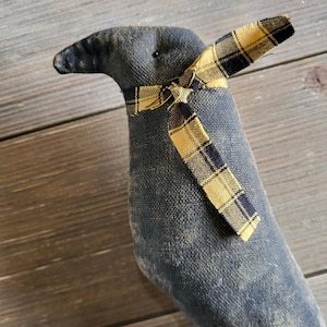 May include: A handmade, stuffed crow doll with a dark gray body and a yellow and black plaid bow tie. The crow has a slightly distressed appearance, with visible stitching and a textured fabric. The background is a wooden surface.