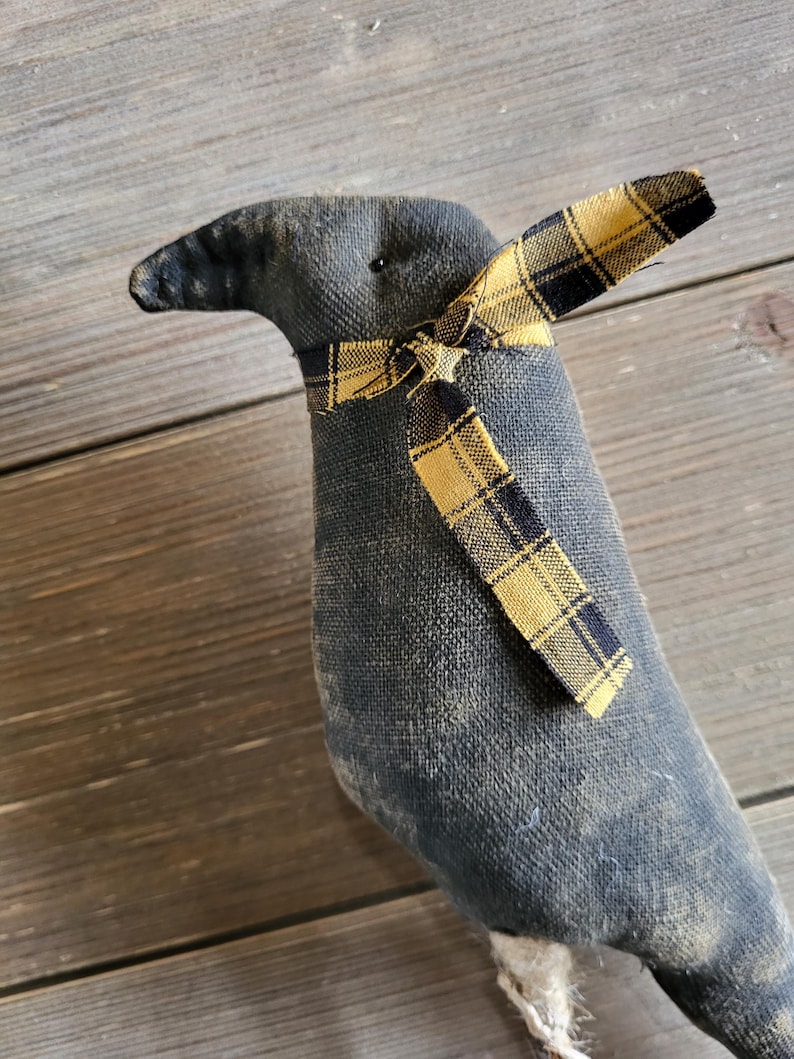 May include: A handmade, stuffed crow doll with a dark gray body and a yellow and black plaid bow tie. The crow has a slightly distressed appearance, with visible stitching and a textured fabric. The background is a wooden surface.