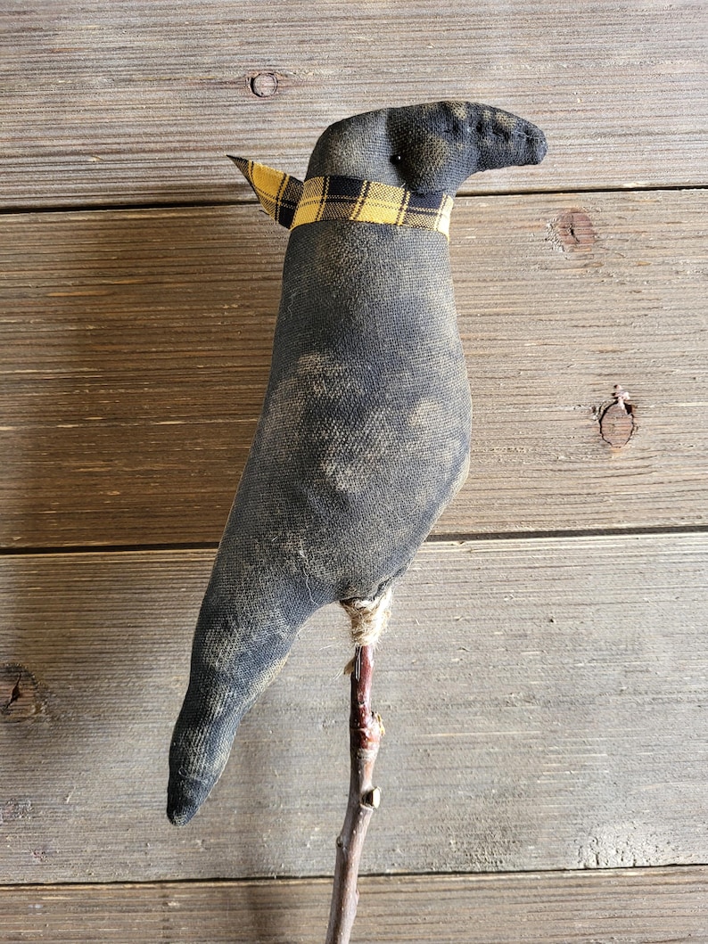 May include: A handcrafted black fabric crow figurine with a yellow and black plaid scarf around its neck. The crow is perched on a small wooden branch, creating a rustic, farmhouse aesthetic. The crow's body has a distressed, vintage look.