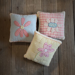May include: Three decorative square pillows with floral designs. One pillow has blue and white stripes with a pink flower. Another has a pink gingham pattern with a white flower. The third pillow is yellow with a pink flower.