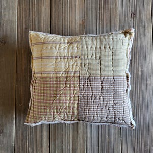 May include: Square patchwork pillow with a variety of patterns in shades of yellow, green, and brown. The pillow features stripes, checks, and solid sections, all stitched together. The edges have a frayed, textured trim, giving it a rustic, handmade appearance.