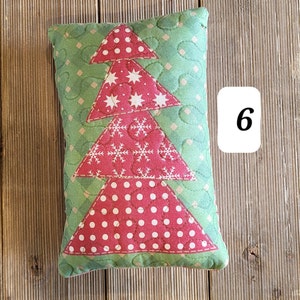 K&ouml;nnte beinhalten: Ein gr&uuml;nes und rotes gestepptes Kissen in Form eines Weihnachtsbaums. Der Baum hat wei&szlig;e Punkte, Sterne und Schneeflocken. Das Kissen liegt auf einer Holzoberfl&auml;che.