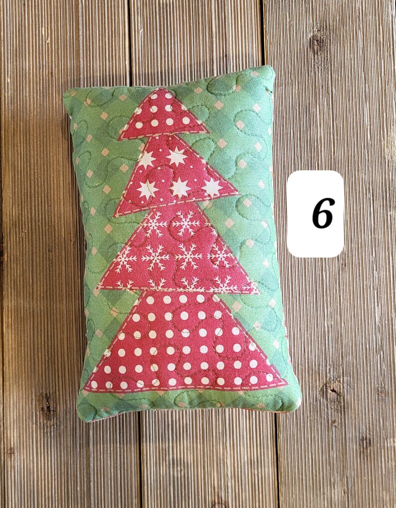 K&ouml;nnte beinhalten: Ein gr&uuml;nes und rotes gestepptes Kissen in Form eines Weihnachtsbaums. Der Baum hat wei&szlig;e Punkte, Sterne und Schneeflocken. Das Kissen liegt auf einer Holzoberfl&auml;che.