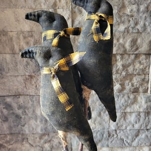 May include: Three black fabric bird figures with long beaks and yellow plaid scarves. The birds are perched on wooden sticks against a textured gray stone wall. The fabric has a distressed, vintage appearance.