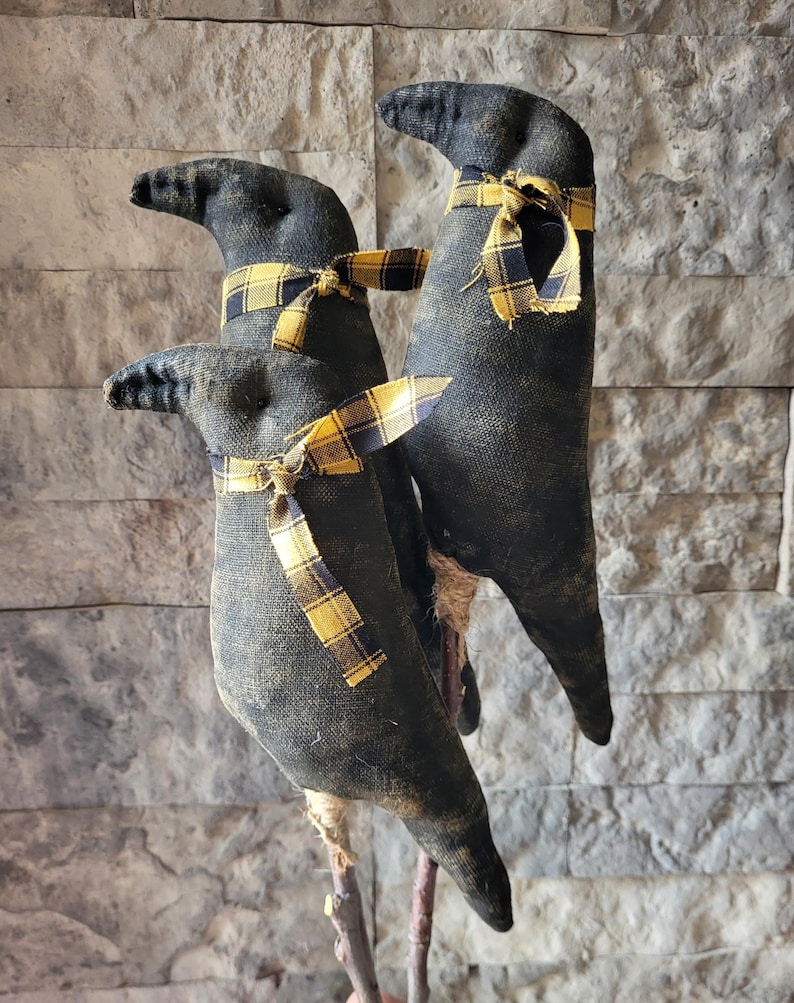 May include: Three black fabric bird figures with long beaks and yellow plaid scarves. The birds are perched on wooden sticks against a textured gray stone wall. The fabric has a distressed, vintage appearance.