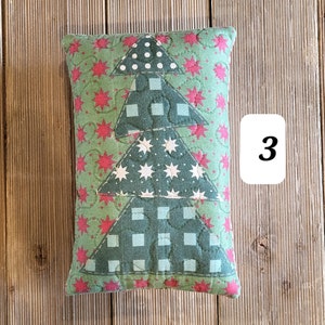 K&ouml;nnte beinhalten: Ein gr&uuml;nes und rotes gestepptes Kissen mit einem Patchwork-Design eines Weihnachtsbaums. Der Baum besteht aus verschiedenen Stoffen mit wei&szlig;en Sternen, roten Sternen und wei&szlig;en Punkten. Das Kissen liegt auf einer Holzoberfl&auml;che.