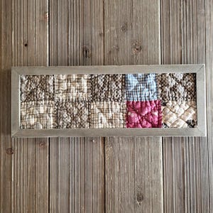 May include: A rectangular wall art piece featuring a quilted fabric panel with a grid of ten squares in shades of brown, beige, blue, and pink, encased in a simple gray wooden frame. The fabric squares display various patterns including checks.