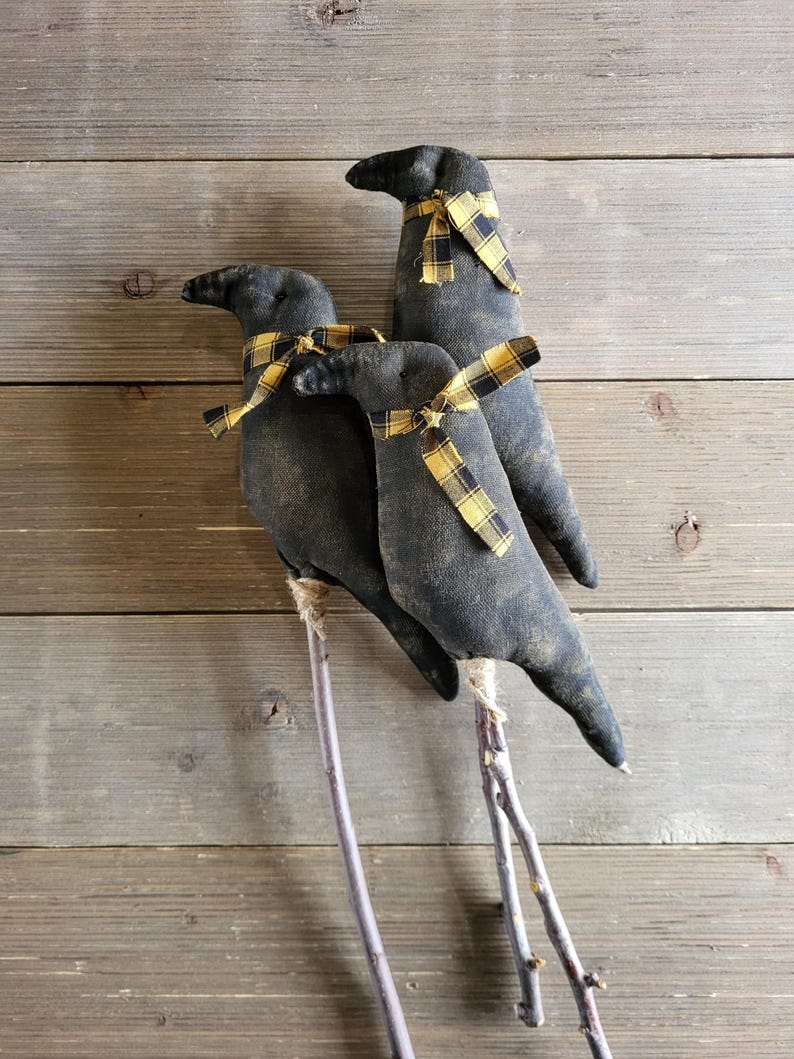 May include: Three black fabric crow decorations with yellow and black plaid ribbon around their necks. The crows are perched on thin, twig-like legs, against a rustic wooden background. A charming, handcrafted, seasonal decoration.