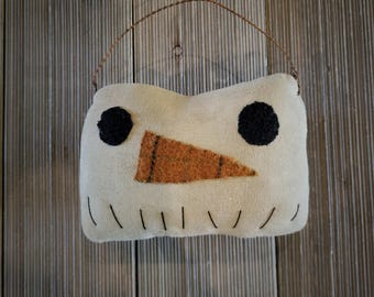 May include: A snowman-shaped decorative pillow with a cream-colored body, black eyes, and an orange triangle nose. The pillow has a brown wire loop for hanging and is stitched with black thread. The background is a wooden wall.