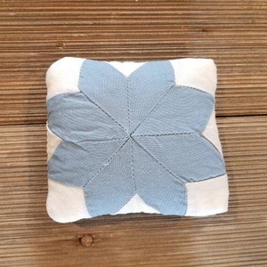 May include: A small, square, blue and white pillow with a star-shaped design. The star is made of blue fabric and is surrounded by white fabric.