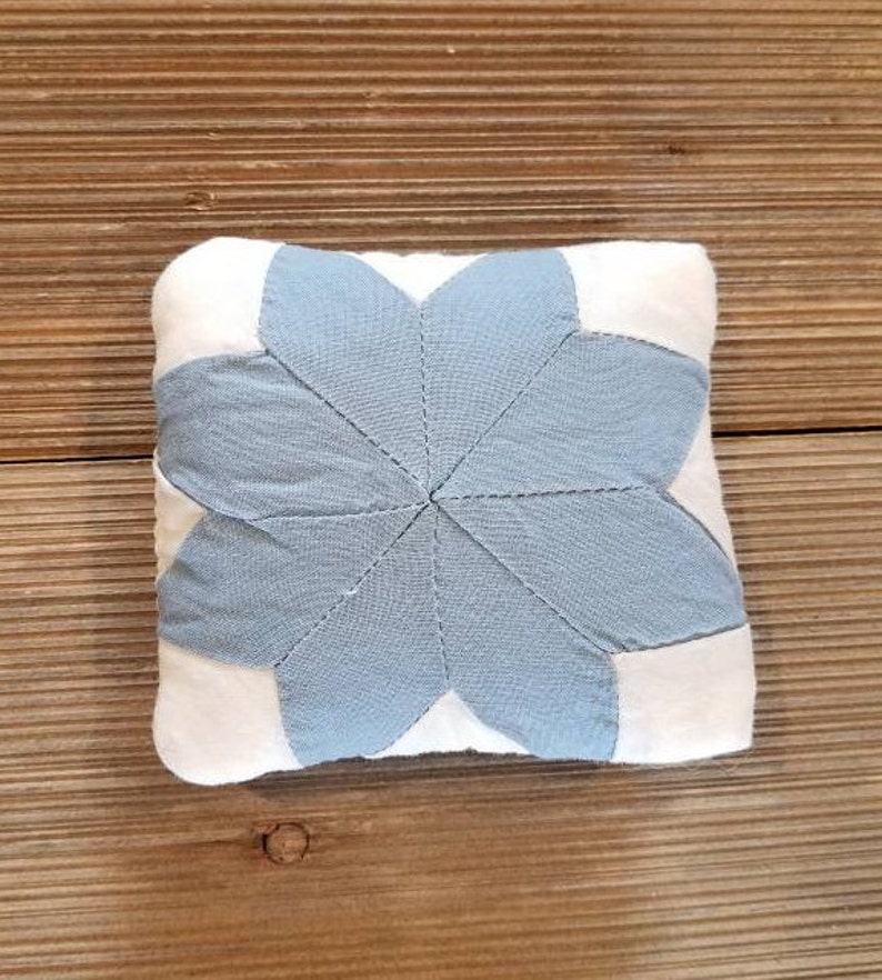 May include: A small, square, blue and white pillow with a star-shaped design. The star is made of blue fabric and is surrounded by white fabric.