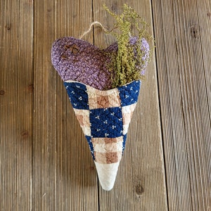 May include: A purple and blue patchwork heart-shaped fabric cone with dried flowers inside. The cone is hanging from a string on a wooden background.