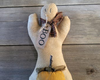 May include: A beige fabric ghost figurine with stitched eyes, wearing a brown plaid scarf and a tag printed with the word BOO. The ghost holds a small, quilted mustard yellow pumpkin with a dark green fabric leaf and a twig stem at its base.