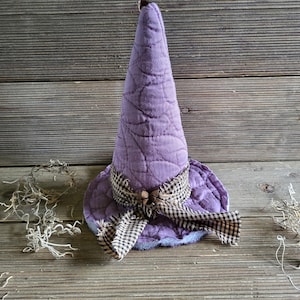 May include: A quilted purple witch hat featuring a tall, pointed crown and a wide brim. The hat is accented with a brown and cream checkered fabric ribbon tied in a bow at the base, which is decorated with two small, round, light brown wooden beads.