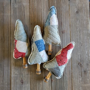 May include: Four handmade fabric Christmas tree ornaments. Each tree is a patchwork of red, white, blue, and gray fabrics, with a quilted texture. The trees are mounted on wooden dowels with silver-colored metal accents.