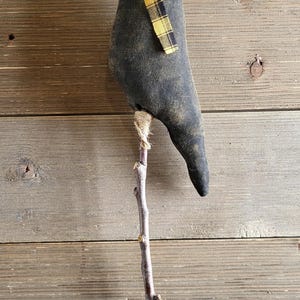 May include: A handcrafted black fabric crow figurine with a yellow and black plaid scarf, perched on a natural wooden stick. The crow has a rustic, homespun aesthetic, perfect for fall or Halloween decor.