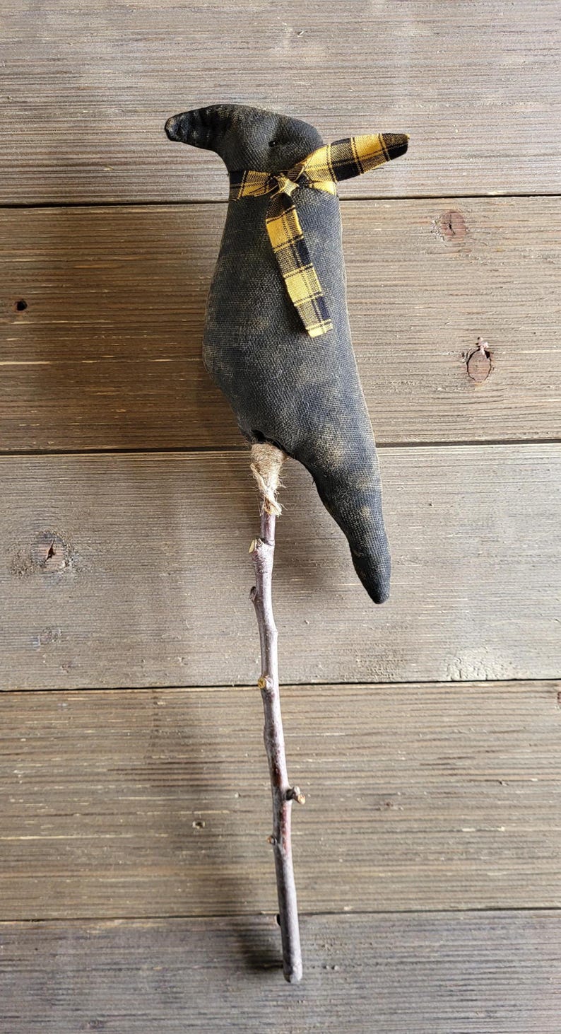 May include: A handcrafted black fabric crow figurine with a yellow and black plaid scarf, perched on a natural wooden stick. The crow has a rustic, homespun aesthetic, perfect for fall or Halloween decor.