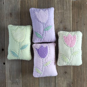 May include: Four rectangular decorative pillows with tulip designs. The pillows are in shades of green, lavender, and pink, with green and lavender stitched details. The pillows are arranged on a wooden surface.