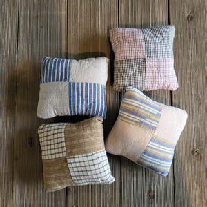 May include: Four small, square decorative pillows with patchwork designs. The pillows feature various patterns, including stripes and plaid, in shades of blue, pink, beige, and brown. They are arranged on a rustic wooden surface.