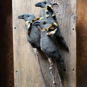 May include: Three dark gray fabric crow figures with twig legs and yellow and black plaid ribbon scarves. The crows are mounted on a weathered wooden board, creating a rustic, country-style decorative piece.