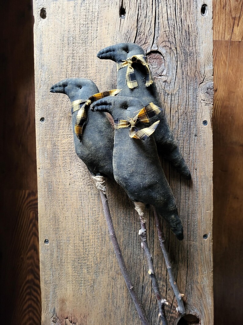 May include: Three dark gray fabric crow figures with twig legs and yellow and black plaid ribbon scarves. The crows are mounted on a weathered wooden board, creating a rustic, country-style decorative piece.