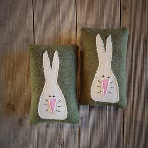 May include: Two green felt bunny pillows with white bunny faces and pink noses. The pillows are on a wood background.