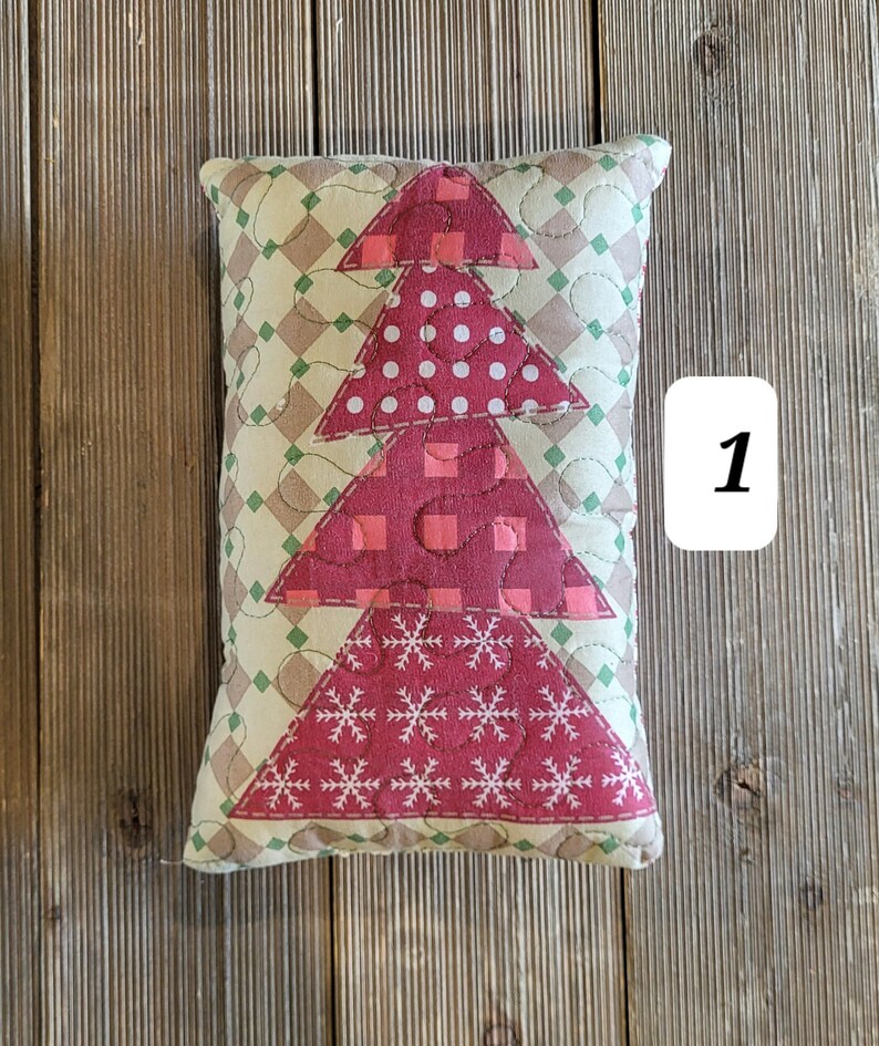 K&ouml;nnte beinhalten: Ein rotes, gr&uuml;nes und wei&szlig;es gestepptes Kissen mit einem Weihnachtsbaum-Motiv. Das Kissen liegt auf einer Holzoberfl&auml;che.