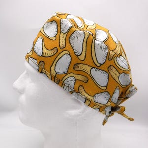 May include: A yellow surgical scrub cap with a repeating pattern of white and tan shapes. The cap is tied at the back and is displayed on a white mannequin head. The fabric has a soft, matte finish.