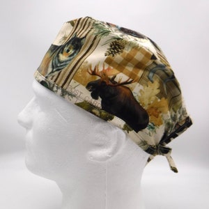 Puede incluir: Un gorro quirúrgico estampado con un alce, un lobo y otras ilustraciones con temática de la naturaleza. El gorro está hecho de una tela de color claro con un diseño repetido de vida silvestre y follaje. Las palabras "POST CARD" son visibles en el gorro.