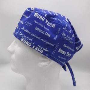 Könnte beinhalten: Eine blaue OP-Haube mit weißem Text. Die Haube zeigt Sätze wie "SURG TECH", "Surgical Care" und "Scrubs". Die Haube wird am Rücken gebunden und wird auf einem weißen Schaufensterkopf präsentiert.