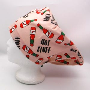 May include: A light pink chef's hat with a repeating pattern of red and green hot sauce bottles. The words "HOT STUFF" are printed in black on the hat.