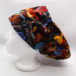 Può includere: Un cappello chirurgico nero con un motivo colorato di chitarre elettriche e acustiche in vari colori. Il cappello è esposto su una testa di manichino bianca, mettendo in risalto il design della chitarra.
