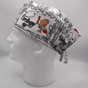 May include: White surgical scrub cap with a repeating chain pattern in gray. The cap features the word "Urology" in black script and a cartoon character holding a wrench and toolbox. The cap is tied at the back.
