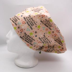 May include: A peach-colored surgical scrub cap with a repeating pattern of colorful stars and the text "You don't have to be crazy to work here... We'll train you." The cap is designed to cover the head and hair.
