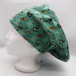 May include: A green scrub cap with a repeating pattern of chickens, blue flowers, and the text "FLUENT IN FOWL LANGUAGE". The cap is made of fabric and has a drawstring closure.