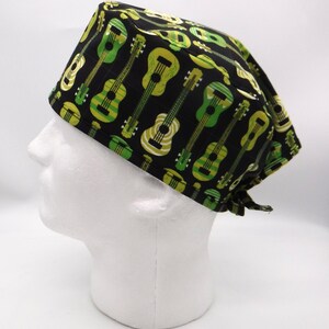 Può includere: Un cappello chirurgico nero con un motivo ripetuto di chitarre verdi e gialle. Le chitarre variano nello stile e nella tonalità, creando un tema musicale. Il cappello è legato sul retro.