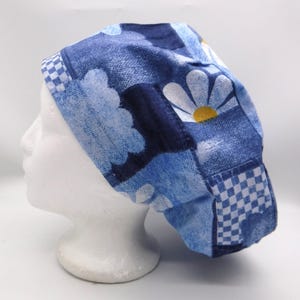 Könnte beinhalten: Eine blau-weiß gemusterte OP-Haube mit einem Gänseblümchen- und Wolkenmuster. Die Haube ist aus Stoff gefertigt und hat ein Schachbrettmuster am Rand. Die Haube wird auf einem weißen Schaufensterkopf präsentiert.