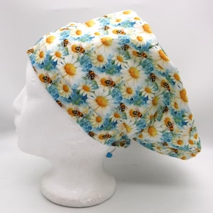 Peut inclure: Une coiffe de chirurgien à motif floral blanc et jaune avec des marguerites et des abeilles. Le bonnet est posé sur un fond blanc et est conçu pour être porté sur la tête. Il est orné d'un cordon de serrage bleu.
