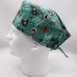 May include: A teal surgical scrub cap featuring a pattern of cartoon chickens, blue flowers, and the text "FLUENT IN FOWL LANGUAGE." The cap is tied at the back and is designed to be worn during medical procedures.